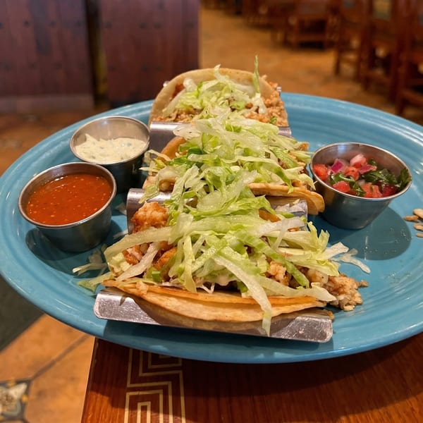 Three tacos filled with meat and shredded lettuce, served with salsa and sauce on a blue plate.