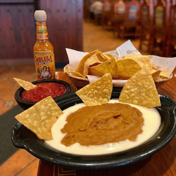 A Cholula hot sauce bottle, salsa, queso, refried beans, and tortilla chips on a wooden table.