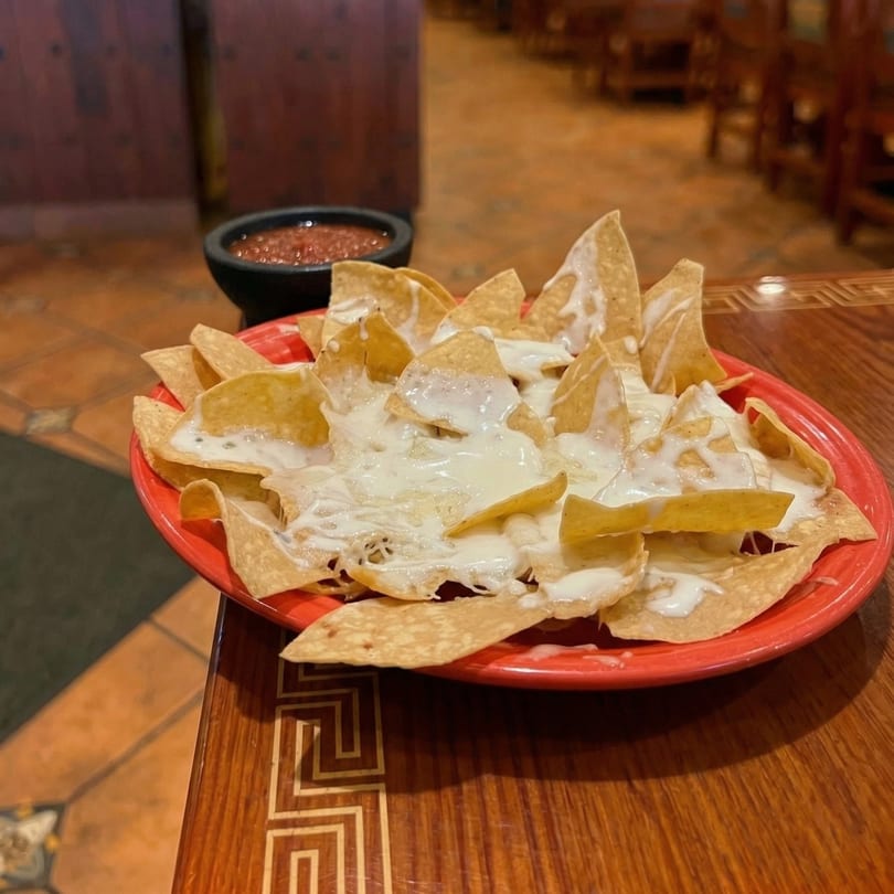 A red plate of cheesy nachos with a side of salsa on a wooden table in a restaurant.