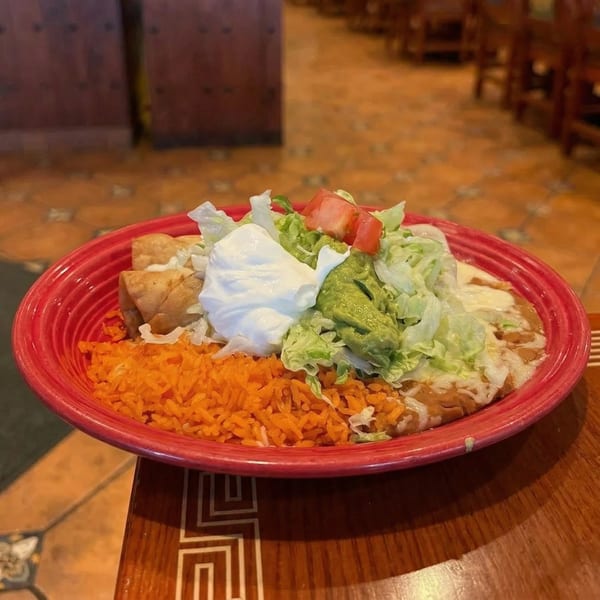 A red plate of Mexican food with taquitos, rice, beans, lettuce, tomatoes, guacamole, and sour cream.