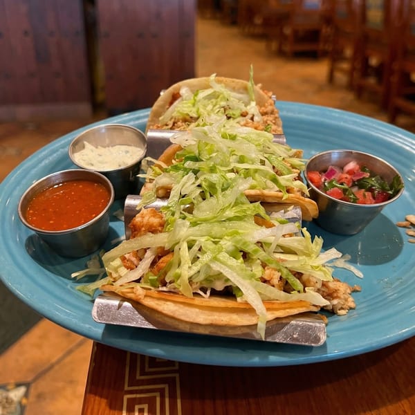 Three tacos with lettuce and meat, served with salsa, pico de gallo, and crema on a blue plate.
