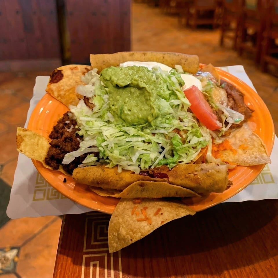 An orange plate filled with various Mexican dishes, including tacos, taquitos, and nachos, topped with guacamole and shredded lettuce.
