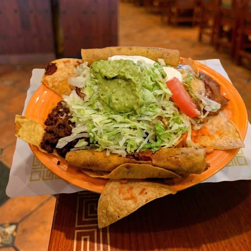 An orange plate filled with various Mexican dishes, including tacos, taquitos, and nachos, topped with guacamole and shredded lettuce.