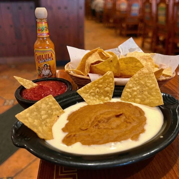 A plate of queso and refried beans with tortilla chips, salsa, and a bottle of Cholula hot sauce.