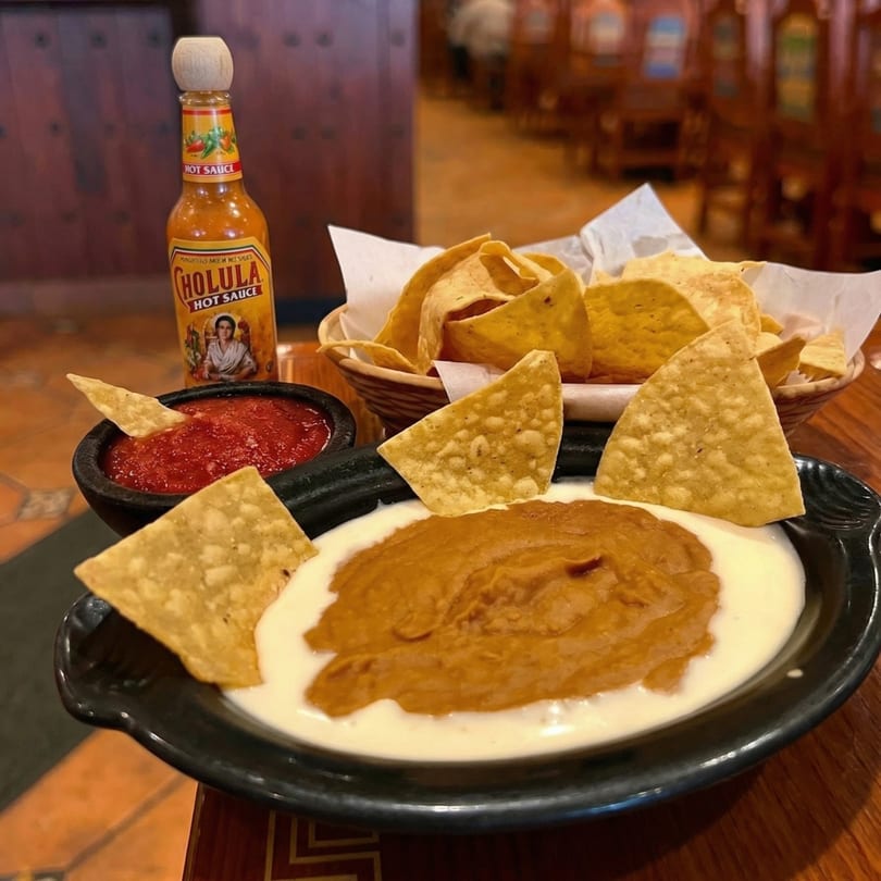 A plate of queso and refried beans with tortilla chips, salsa, and a bottle of Cholula hot sauce.
