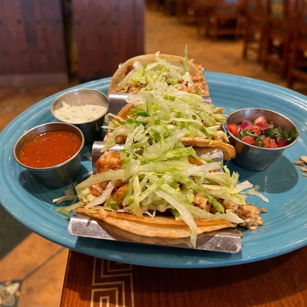 Three tacos filled with meat and shredded lettuce, served with salsa, pico de gallo, and crema.
