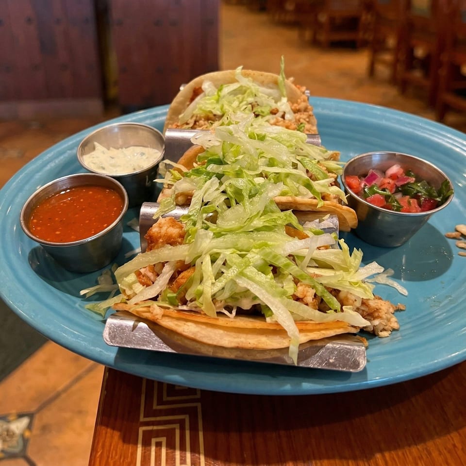 Three tacos with shredded lettuce, meat, and sauces on a blue plate in a restaurant setting.