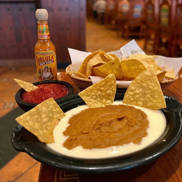 A Cholula hot sauce bottle, a basket of tortilla chips, salsa, and a queso dip.